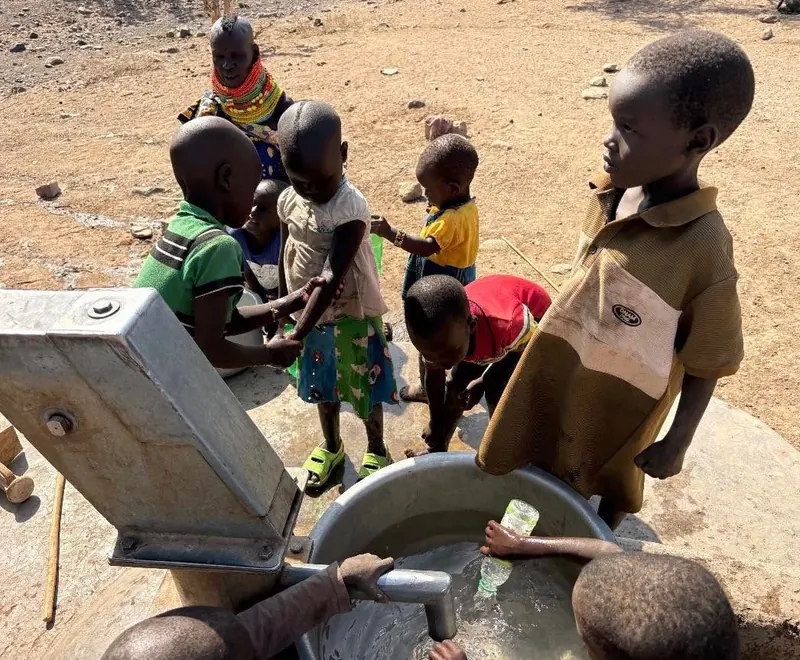 Children gather at a hand pump in Kangikukas as water pours into a metal basin, with a woman in beaded necklaces behind them.
