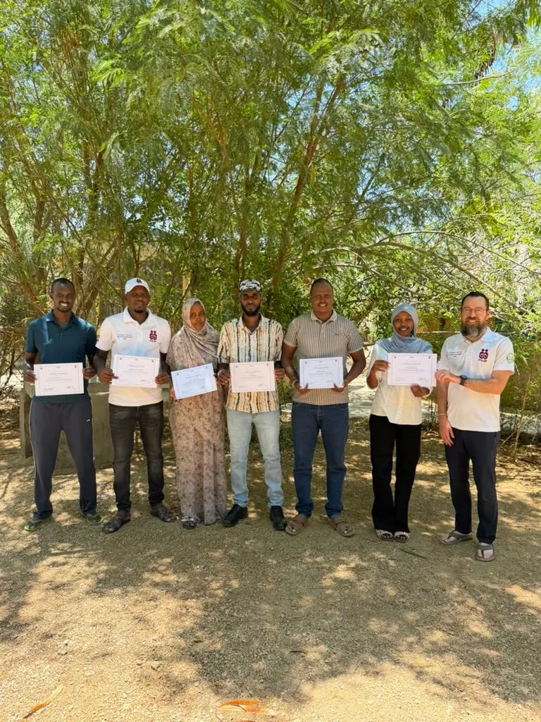 Seven members of the Marsabit COHI team stand under a tree holding their training certificates.